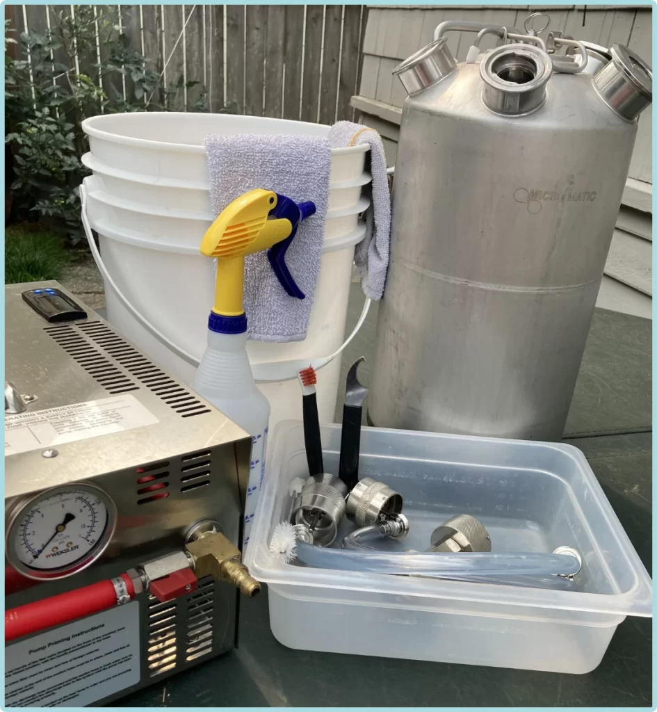 A brewing setup in Tacoma with a large stainless steel keg, stacked white buckets, a spray bottle, towel, sanitizer, hoses, tap line, valves, and various metal brewing tools arranged on a table.