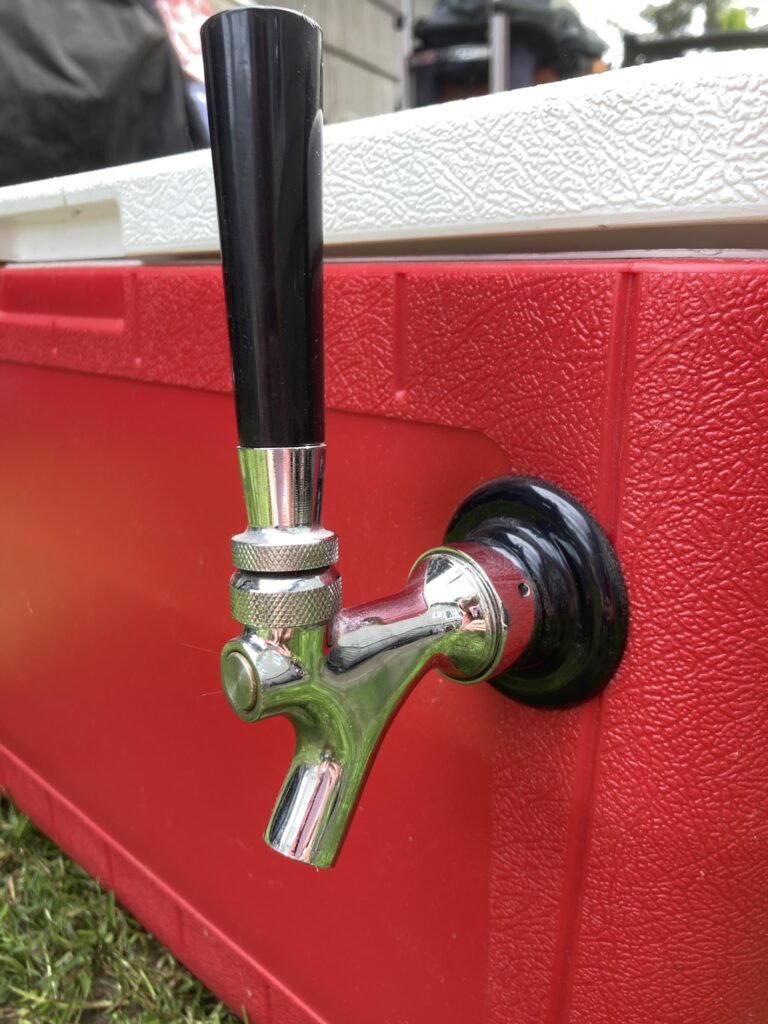 A metal beer tap faucet with a black handle is installed on the side of a red cooler sitting outdoors on grass.