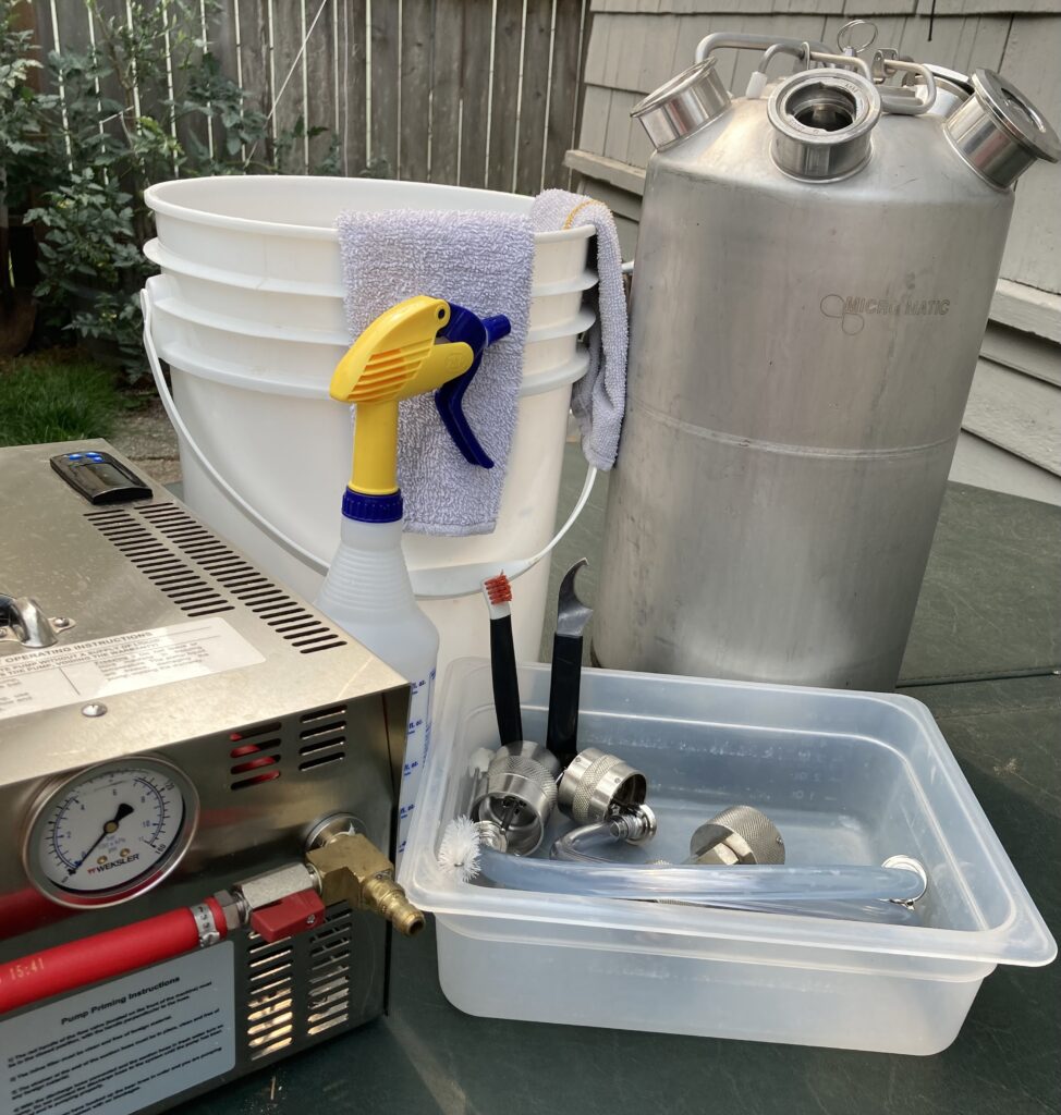 Assorted brewing equipment, including a pressure gauge machine, metal keg, buckets, spray bottle, cleaning towel, and tap line fittings arranged on an outdoor table.