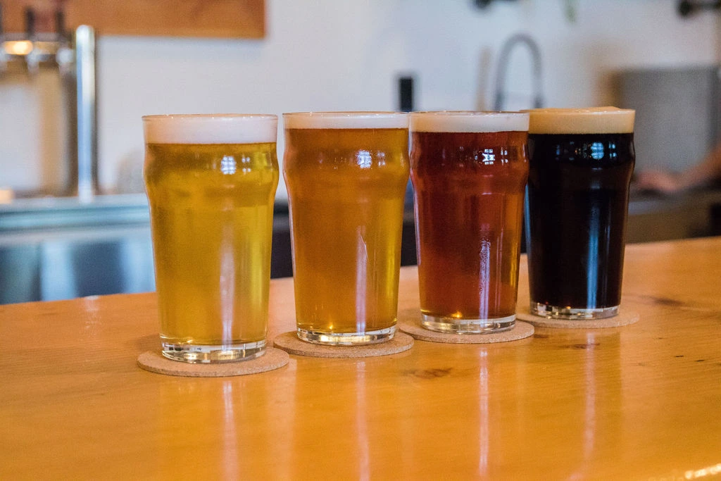 Four pints of beer in different shades from light to dark are lined up on a wooden bar, each glass set on a round coaster, fresh from the tap line and ready to enjoy near Puget Sound.