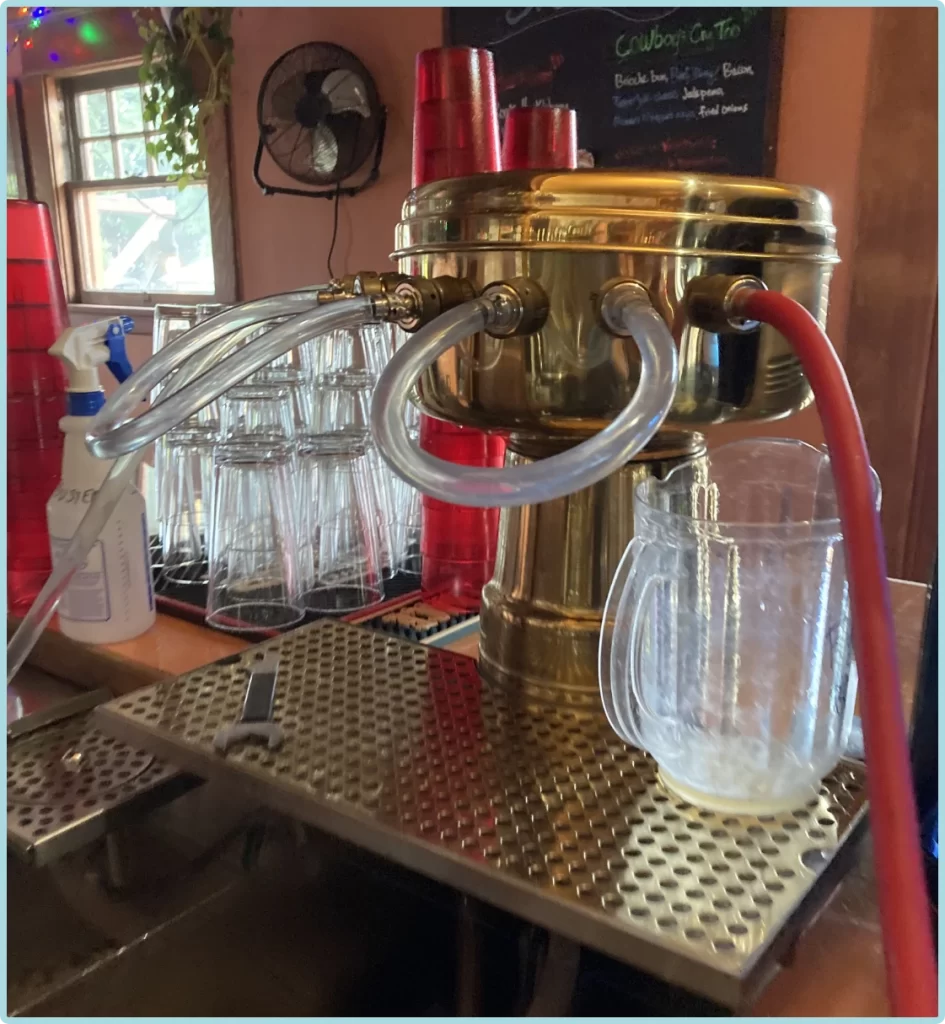 A brass draft beer dispenser with attached tap lines sits on a bar counter near stacked glasses, red cups, and a half-filled pitcher—perfect for enjoying fresh pours in Puget Sound bars after thorough tap cleaning.