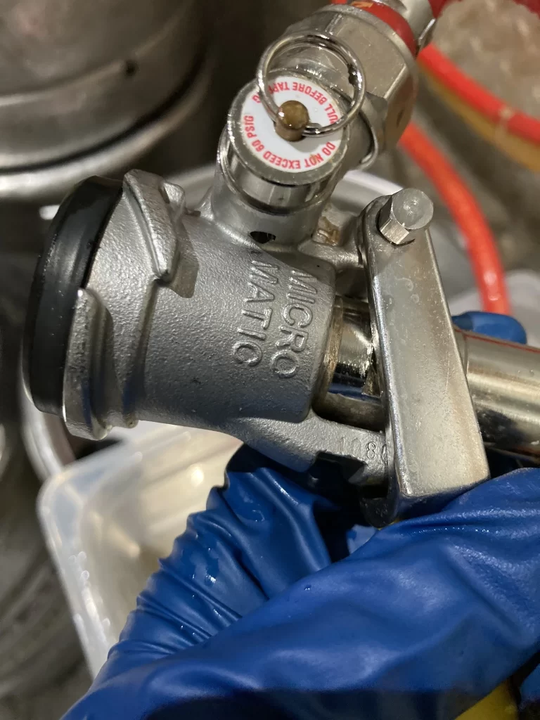 A gloved hand holds a Micro Matic keg coupler connected to a beer keg, with red and silver hoses attached for tap cleaning.