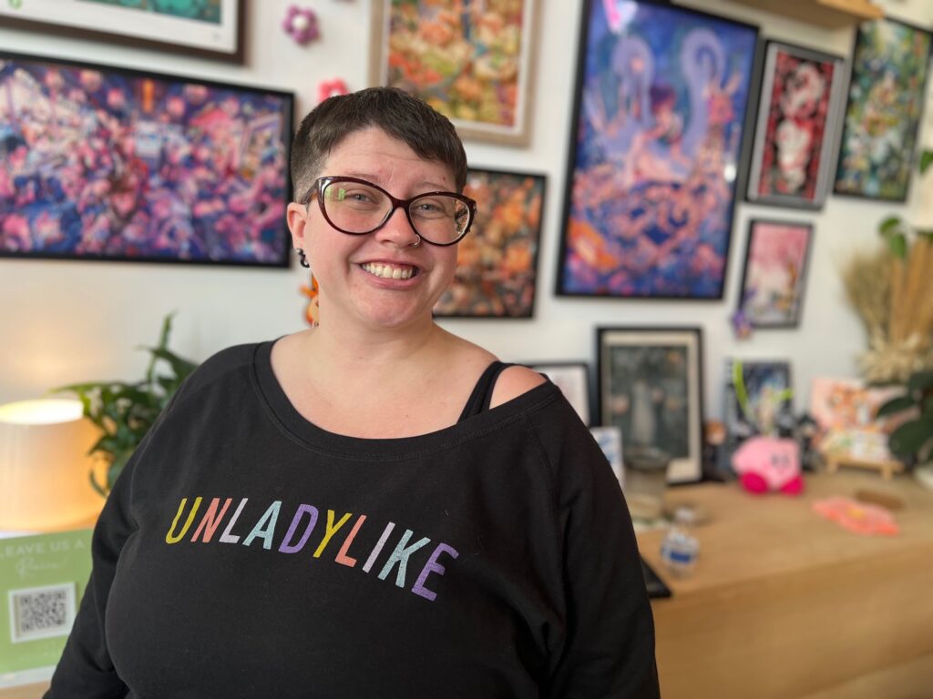A smiling person with short hair and glasses stands indoors, wearing a black shirt that reads "UNLADYLIKE," with colorful artwork hanging on the wall behind them.