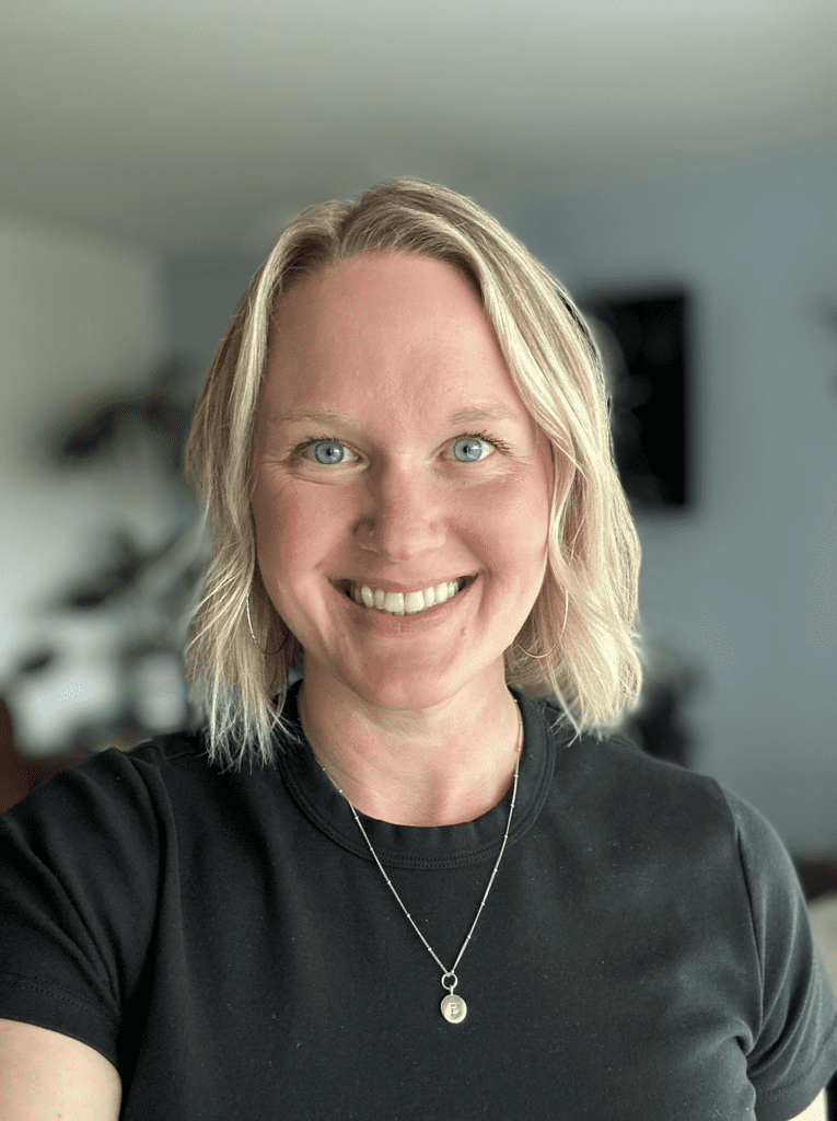 A woman with blonde hair and blue eyes, wearing a black shirt and a necklace, smiles at the camera in a well-lit indoor setting.