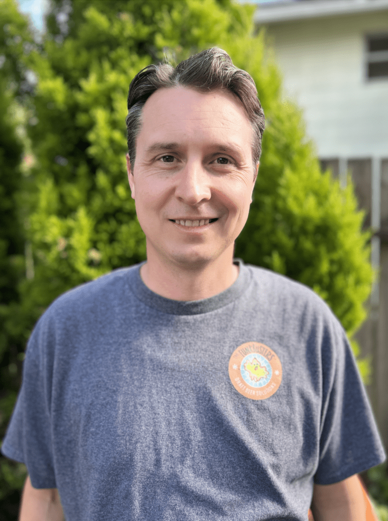 A man with short dark hair smiles at the camera, wearing a gray T-shirt with a circular logo, standing outdoors in front of green bushes and a wooden fence.