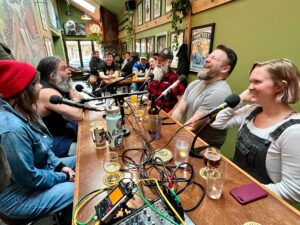 A group of people sit around a long table with microphones and drinks, recording a podcast in a lively pub setting.