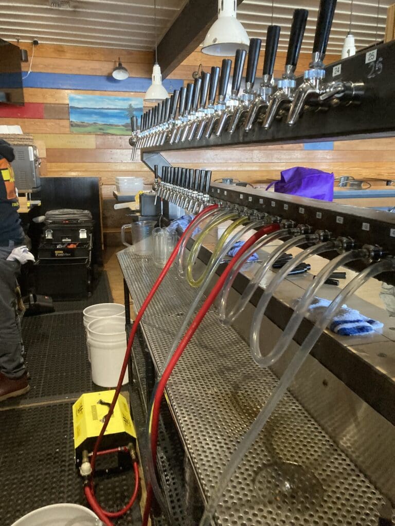 Two horizontal rows of beer taps mounted on a wall with clear tubes running from the lower taps into buckets on the floor.